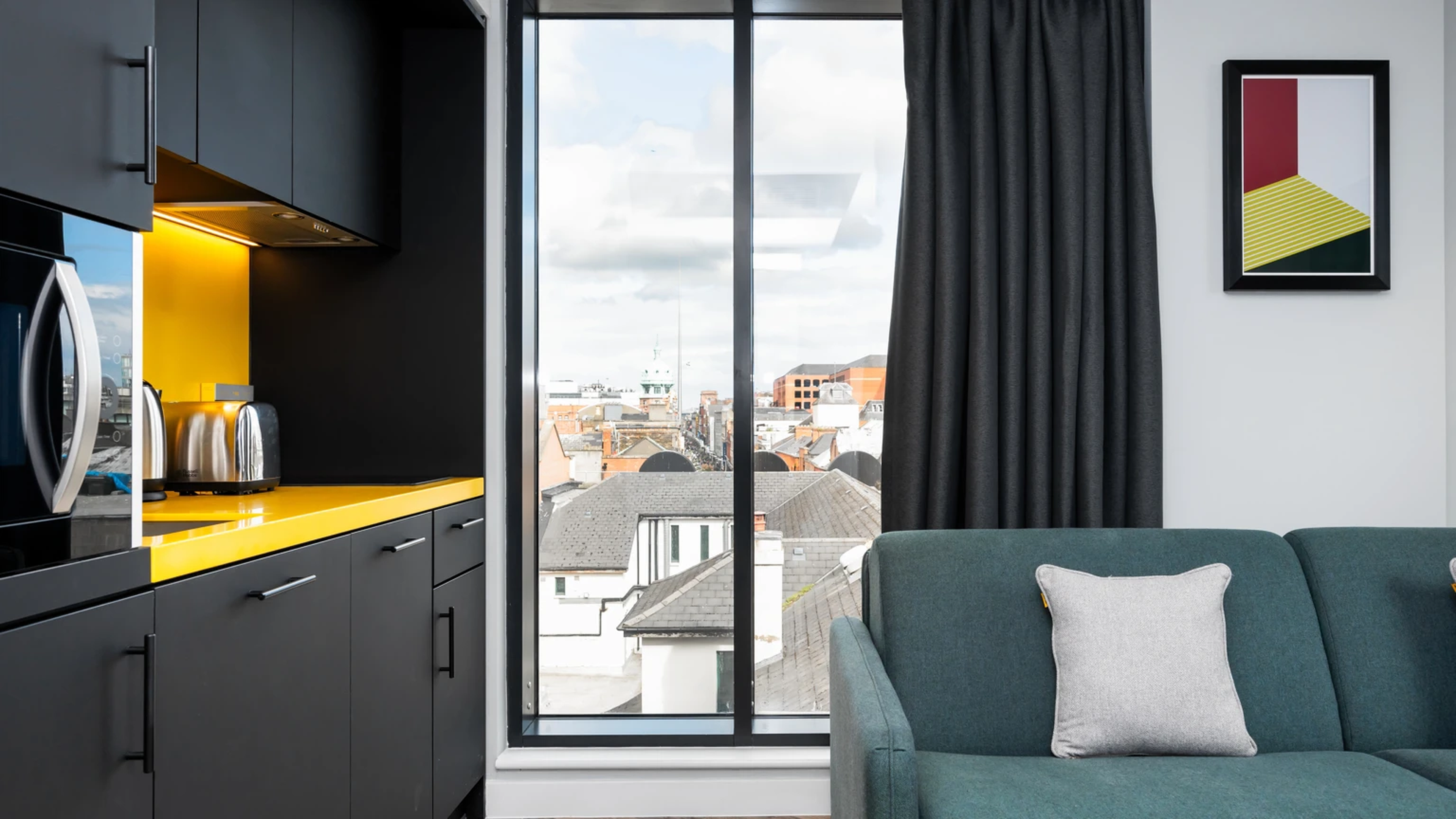 Modern apartment interior with a black and yellow kitchen, large window with city view, teal sofa, and framed geometric art on the wall.
