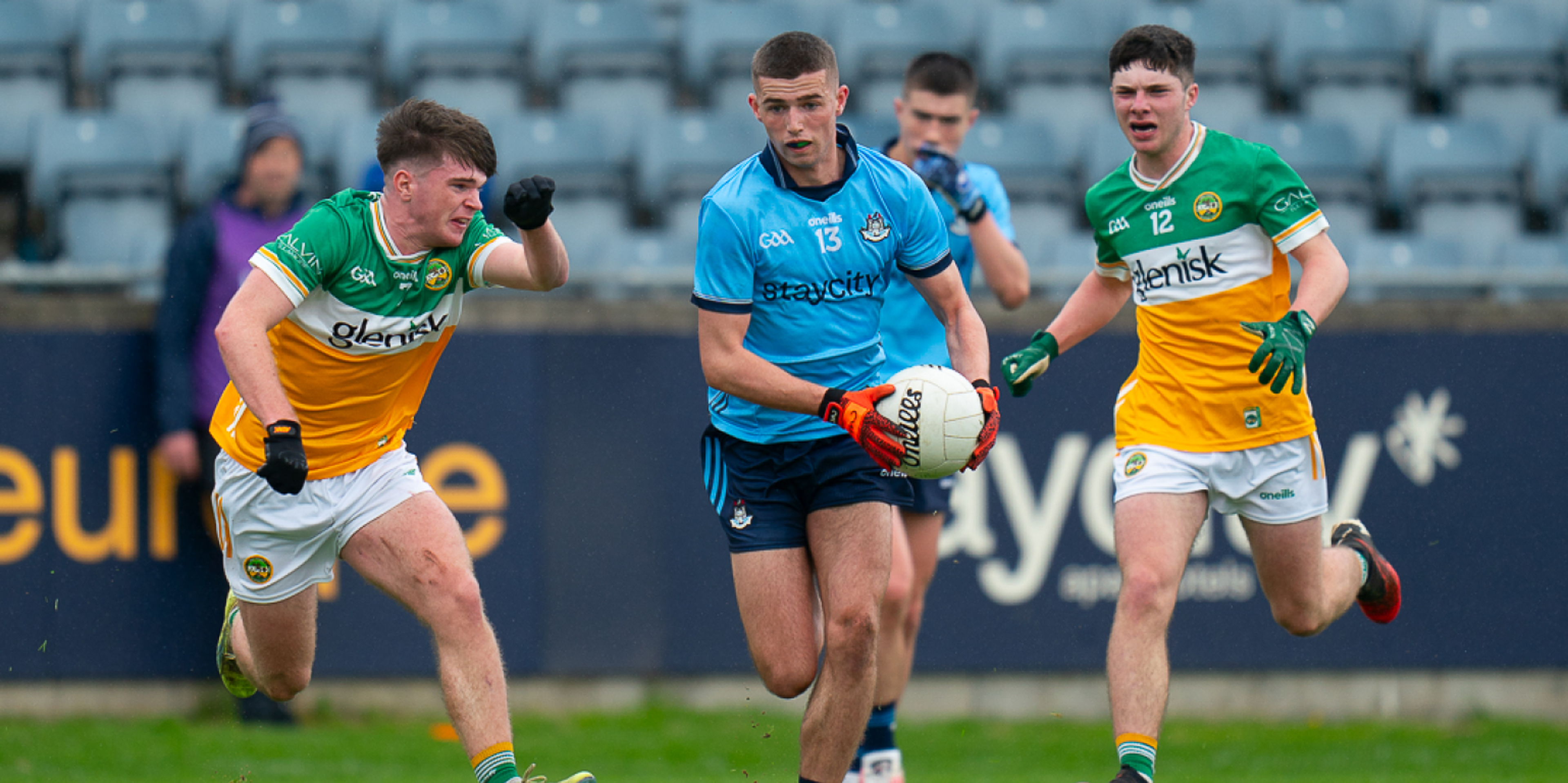Three Gaelic football players in action on the field, one in a blue jersey holding the ball, pursued by two players in green and yellow jerseys.