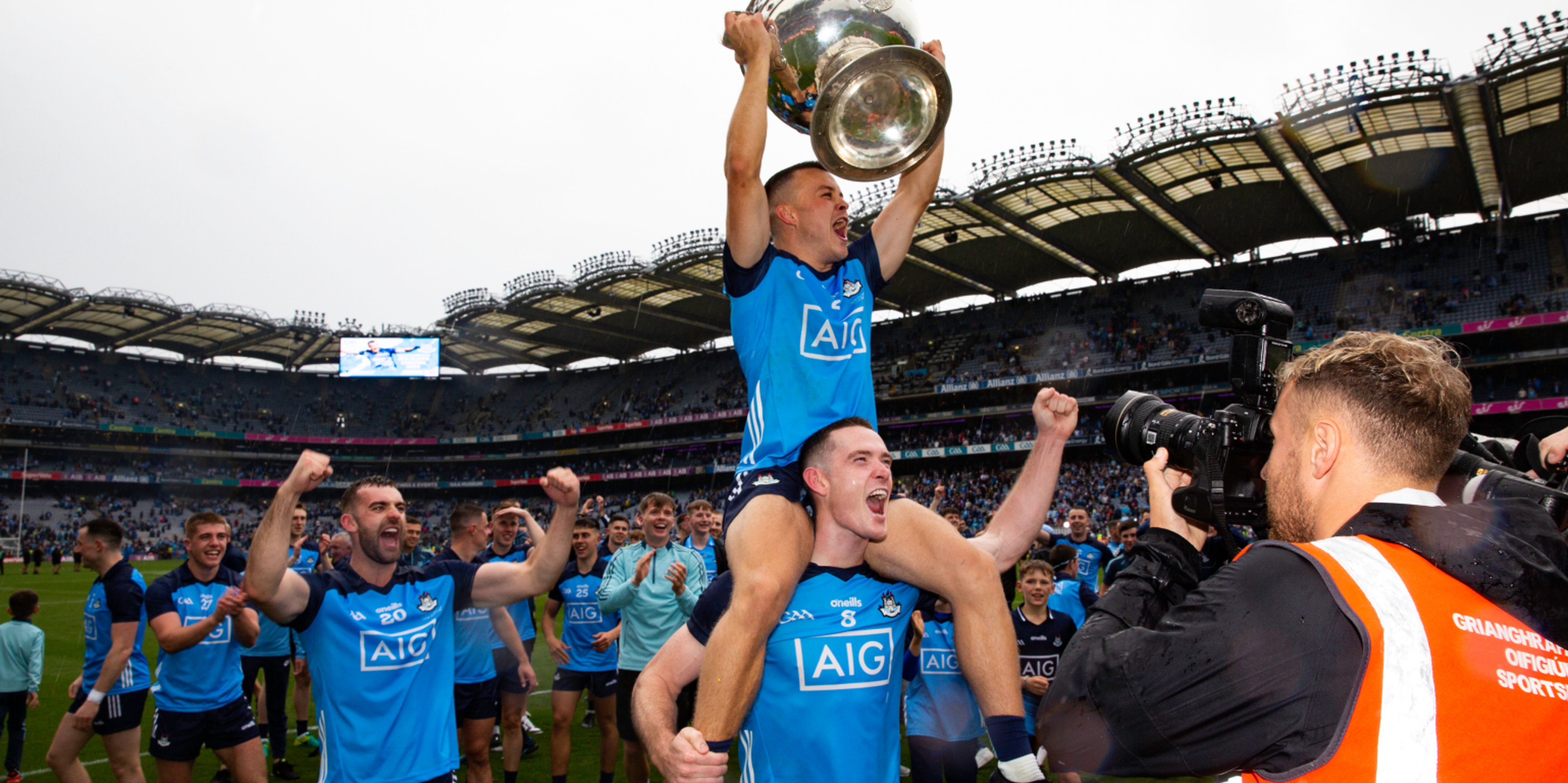 A sports team celebrates on a field; a player lifts a trophy while sitting on a teammate's shoulders, surrounded by cheering teammates and a photographer.
