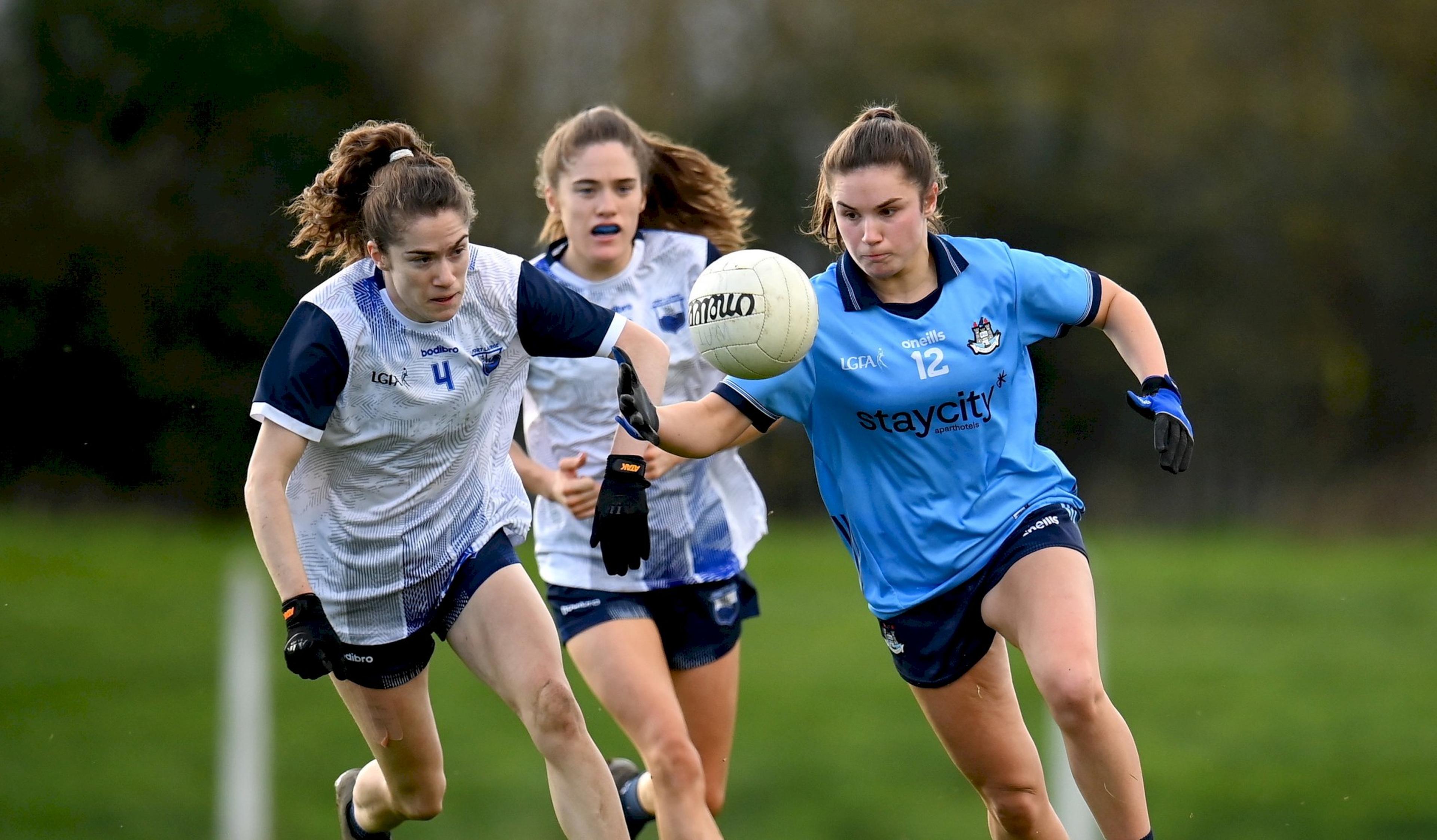 Three women playing Gaelic football on a field; one in a blue jersey reaching for the ball, pursued by two in white jerseys.